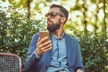 Portrait of a bearded male with a stylish haircut in casual clothes, writes a message on the phone while sitting on a bench in a park.の写真素材