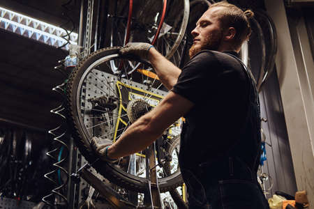 Handsome redhead male in a jeans coverall, working with a bicycle wheel in a repair shop. A worker removes the bicycle tire in a workshop.の写真素材