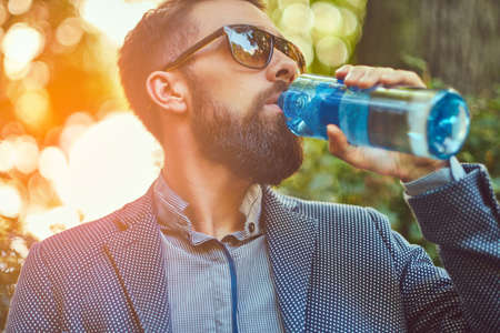 Bearded male man drinking cool water outdoors, sitting on a bench in a city park.の写真素材