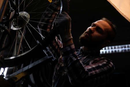 Handsome stylish male wearing a flannel shirt and jeans coverall, working with a bicycle wheel in a repair shop.の写真素材