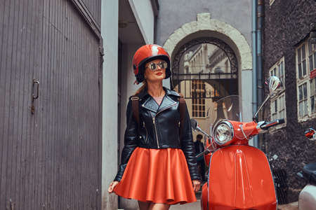 Sexy blonde girl wearing stylish clothes in sunglasses and helmet, standing on an old narrow street with two retro scooters.の写真素材