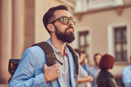 Tourist with a full beard and haircut, wearing casual clothes and sunglasses, holds a backpack, standing on an antique street, during the excursion in Europe.の写真素材
