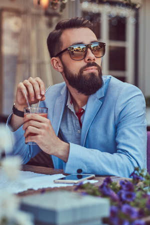 Portrait of a fashionable bearded male with a stylish haircut, holds a glass of a cappuccino, sitting in a cafe outdoors.の写真素材