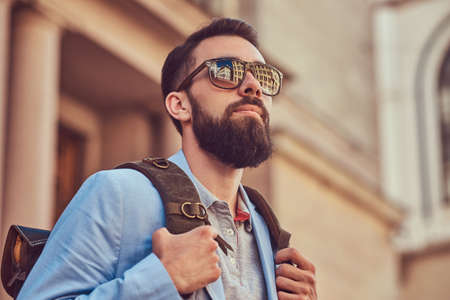 Tourist with a full beard and haircut, wearing casual clothes and sunglasses, holds a backpack, standing on an antique street, during the excursion in Europe.の写真素材