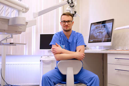 Portrait of a handsome bearded dentist male in glasses wearing a blue uniform, sitting in a dentist office.の写真素材
