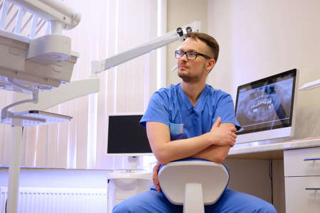 Portrait of a handsome bearded dentist male in glasses wearing a blue uniform, sitting in a dentist office.の写真素材