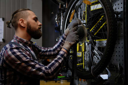 Handsome stylish male wearing a flannel shirt and jeans coverall, working with a bicycle wheel in a repair shop.の写真素材