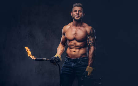 Brutal tattoed male welder with a stylish haircut and beard, with muscular body, dressed in only jeans, holds propane tank and a burning burner, standing in a studio, looking at a camera.の写真素材