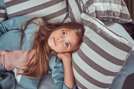 Portrait of a cute little girl with long brown hair and piercing glance, looking at a camera, lying on a sofa at home aloneの写真素材