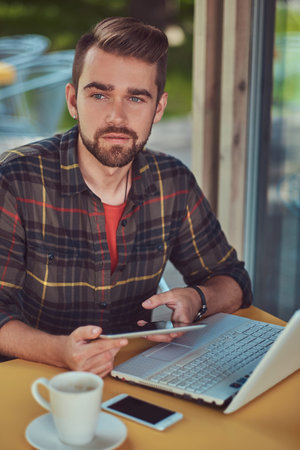 A handsome fashionable male freelancer with stylish haircut and beard, wearing fleece shirt, working...