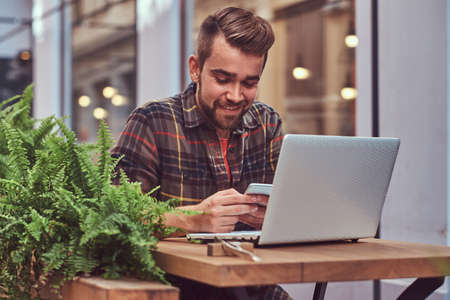 Portrait of a smiling fashionable bearded male with a stylish haircut, dressed in a flannel shirt, sitting in a cafe outdoors.の写真素材