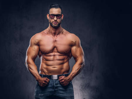 Portrait of a shirtless tall huge male with a muscular body with a stylish haircut and beard, in a sunglasses and jeans, posing in a studio.の写真素材