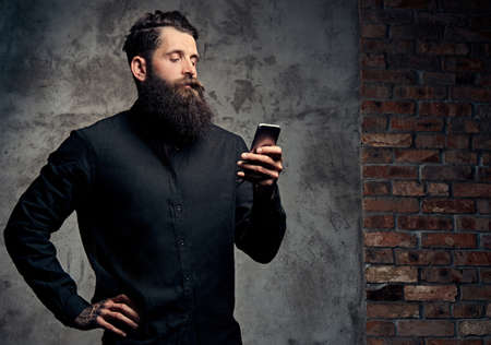 Portrait of a handsome bearded hipster dressed in a black shirt, using a smartphone, standing in a studio, isolated on a dark background.の写真素材