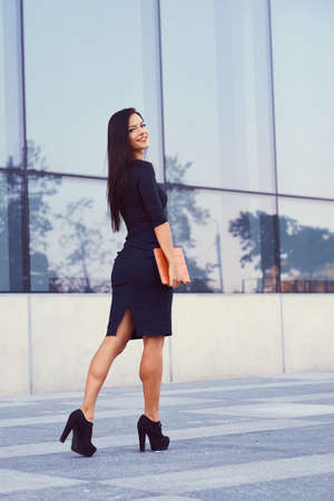 A sexy brunette businesswoman dressed in a black formal clothes and high heels, holds a tablet computer goes to work, against of a skyscrapers.の写真素材
