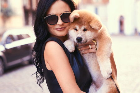 Stylish brunette woman in a black dress and sunglasses, standing on a street and holds a cute purebred puppy Akito.の写真素材