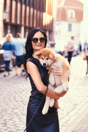 Stylish brunette woman in a black dress and sunglasses, standing on a street and holds a cute purebred puppy Akito.の写真素材