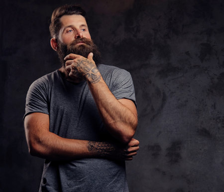 Portrait of a hipster with full beard and stylish haircut, dressed in a gray t-shirt, stands with a...