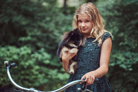 Portrait of a little blonde girl in a casual dress, holds cute spitz dog, in a park.の写真素材