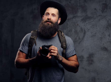 Portrait of a handsome bearded traveler in a hat with a backpack and tattooed arms, holds a photo camera, isolated on a dark background.の写真素材