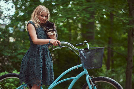 Portrait of a little blonde girl in a casual dress, holds cute spitz dog, in a park.の写真素材