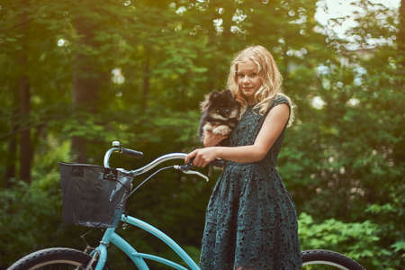 Portrait of a little blonde girl in a casual dress, holds cute spitz dog, in a park.の写真素材