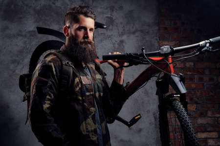 A handsome bearded traveler, dressed in a trendy camouflaged jacket, standing with sports bicycle on...