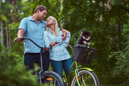 An attractive couple of a blonde female and man dressed in casual clothes on a bicycle ride with their cute little spitz in a bicycle basket.の写真素材