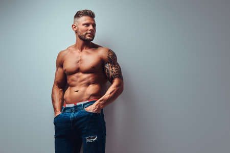 A handsome shirtless tattooed bodybuilder with stylish haircut and beard, wearing sports shorts, posing in a studio. Isolated on a gray backgroundの写真素材
