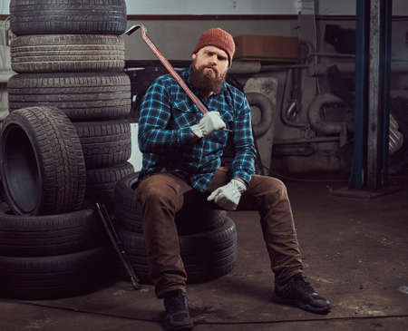A bearded mechanic dressed in a uniform, sits on old car tires in the garage.