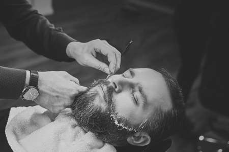 Bearded male sitting in an armchair in a barber shop while hairdresser shaves his beard with a dangerous razor.の写真素材