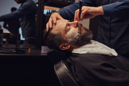 Bearded male sitting in an armchair in a barber shop while hairdresser shaves his beard with a dangerous razor.の写真素材
