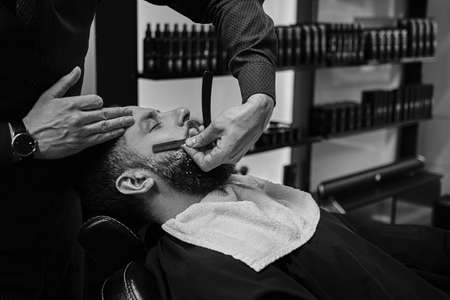 Bearded male sitting in an armchair in a barber shop while hairdresser shaves his beard with a dangerous razor.の写真素材
