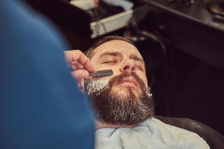 Bearded male sitting in an armchair in a barber shop while hairdresser shaves his beard with a dangerous razor.の写真素材