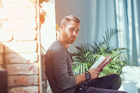 Handsome stylish redhead student in casual clothes, sitting on a chair at a home and holds a book, looking at a camera.の写真素材