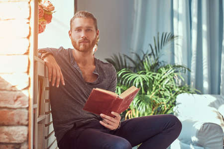 Handsome stylish redhead student in casual clothes, sitting on a chair at a home and holds a book,...