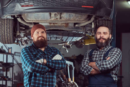 Two bearded brutal mechanics repair a car on a lift in the garage.