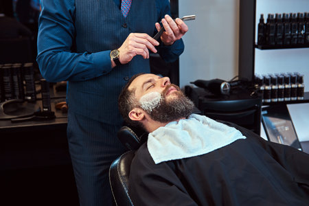 Bearded male sitting in an armchair in a barber shop while hairdresser shaves his beard with a dangerous razor.の写真素材