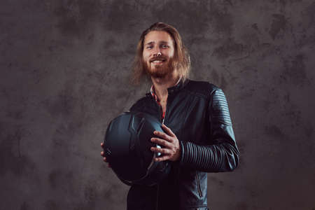 Portrait of a handsome stylish redhead biker in a black leather jacket, holds motorcycle helmet, posing in a studio.の写真素材