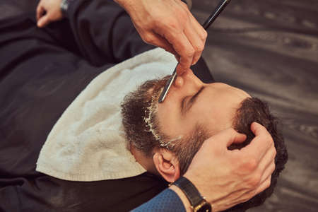 Bearded male sitting in an armchair in a barber shop while hairdresser shaves his beard with a...