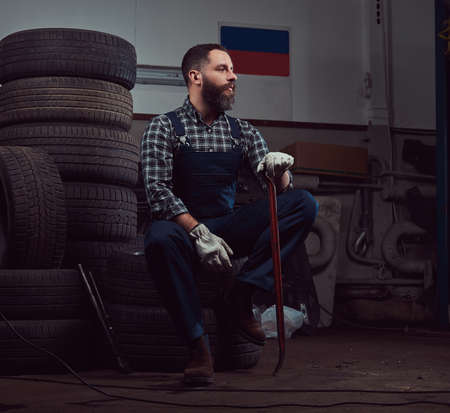 A bearded mechanic dressed in a uniform, sits on old car tires in the garage.の写真素材