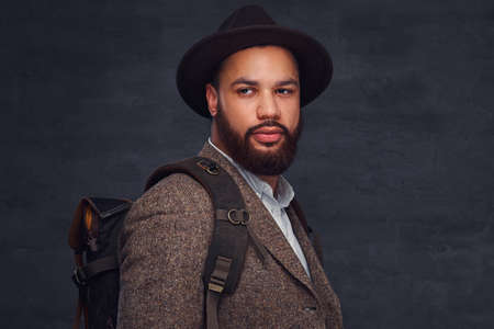 Handsome Afro-American traveler in a brown jacket and hat with the backpack, stands in a studio.の写真素材