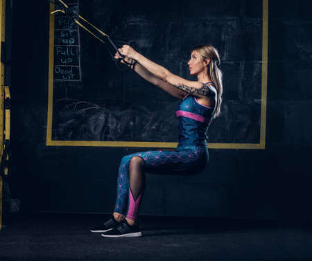Beautiful muscular blonde woman in sportswear with a tattoo on her arm doing exercise with...