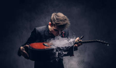 Handsome young musician with stylish hair in elegant clothes exhales smoke on his acoustic guitar.