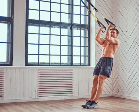 Shirtless muscular male doing exercise with suspensions straps.