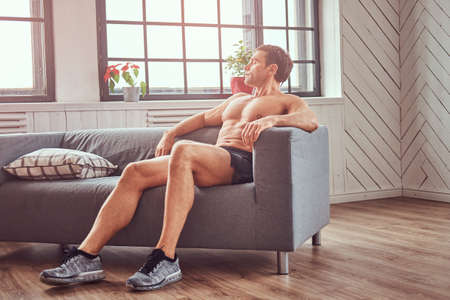 Handsome shirtless muscular man lying on the couch, resting after a hard workout at home.の写真素材