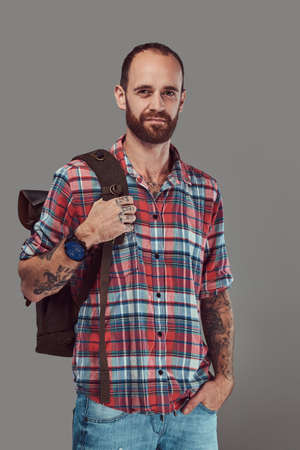 Portrait of a handsome tattooed traveler in a flannel shirt with a backpack, standing in a studio.