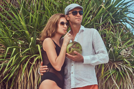 Attractive couple, holds coconut with a tube, posing on a beach near palm bush, enjoys a vacation on a beautiful island.の写真素材