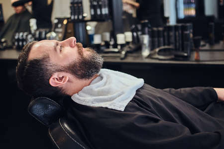 Handsome bearded male waits for the hairdresser sitting on a barber chair.の写真素材