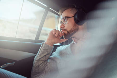 Successful stylish young businessman in a gray suit and pink shirt, riding on a back seat in a luxury car.の写真素材