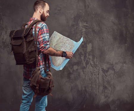Portrait of a handsome tattooed traveler in a flannel shirt with a backpack, holds a map, standing in a studio.の写真素材